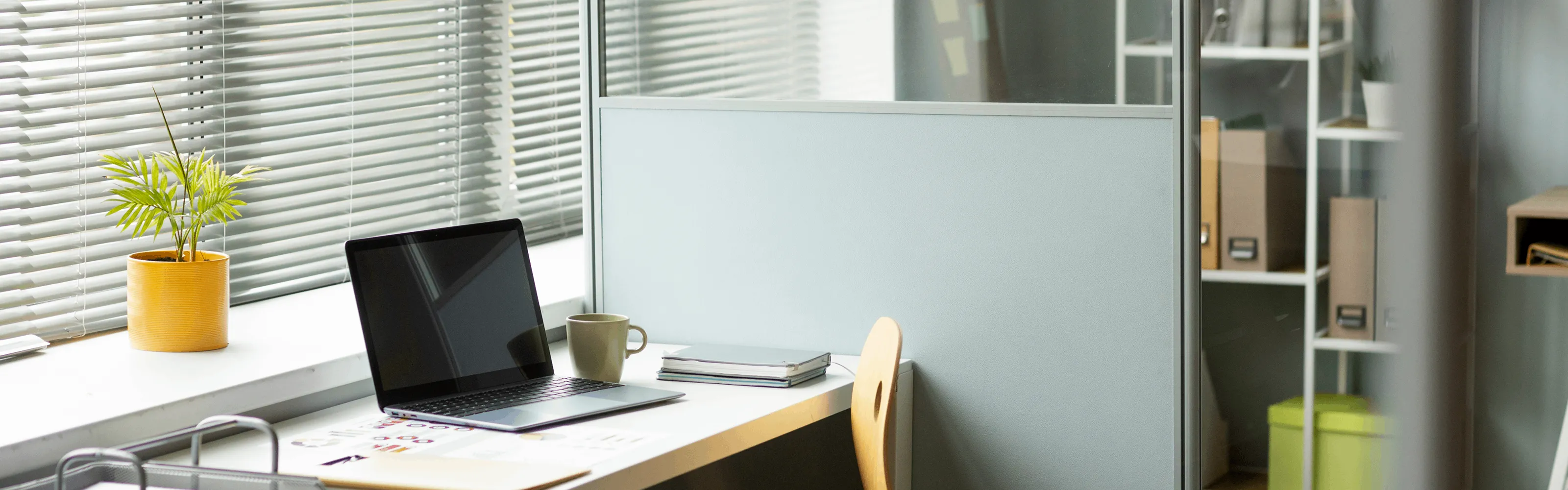 Modern office workspace with laptop, coffee mug, notebooks, and a potted plant by window blinds